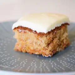 En bit saftig glutenfri morotskaka i långpanna med krämig glasyr på en gråblå tallrik med prickigt mönster.