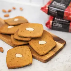 Fler fyrkantiga pepparkakor med mandel ligger på en träskärbräda, med några lösa mandlar och förpackningar i bakgrunden.