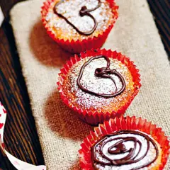 Tre saffransmuffins med mandelmassa, pudrade med florsocker och dekorerade med choklad. En vit sidenband med hjärtan ligger bredvid.