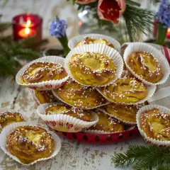 En hög med gyllene kanelbullar, strösslade med pärlsocker, i vita pappersformar. En röd ljusstake och blommor i bakgrunden ger en mysig känsla. Perfekt för att visa hur förvarar man bullar bäst.