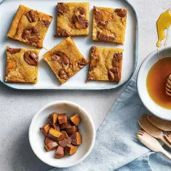 Sju bitar av saftiga dumlekakor i långpanna ligger på ett fat. Bredvid finns en skål med honung och en skål med hackade dumlekolor.