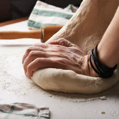 Händer knådar deg på ett mjölat bord. Trots att degen jäser inte, baka ändå! En kavel och en handduk ligger i bakgrunden.