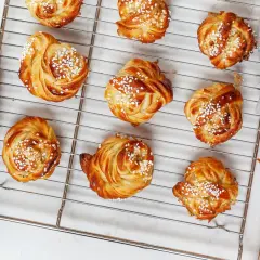 Golden brown cinnamon buns with pearl sugar on a cooling rack, with hands reaching for them.