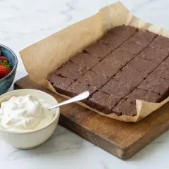 En kladdkaka i långpanna, uppskuren i rutor, serveras med vispad grädde och färska jordgubbar. Perfekt för fikat!