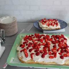 En stor rulltårta som tårta, täckt med grädde och jordgubbar, står på ett grönt fat. En mindre tårta med samma dekoration syns på en blå tallrik.