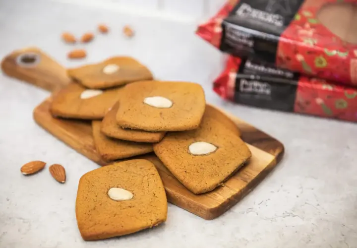 Fler fyrkantiga pepparkakor med mandel ligger på en träskärbräda, med några lösa mandlar och förpackningar i bakgrunden.