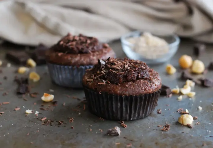 Två chokladmuffins med nutella och strössel av choklad. Hasselnötter och chokladbitar ligger utspridda runt dem.