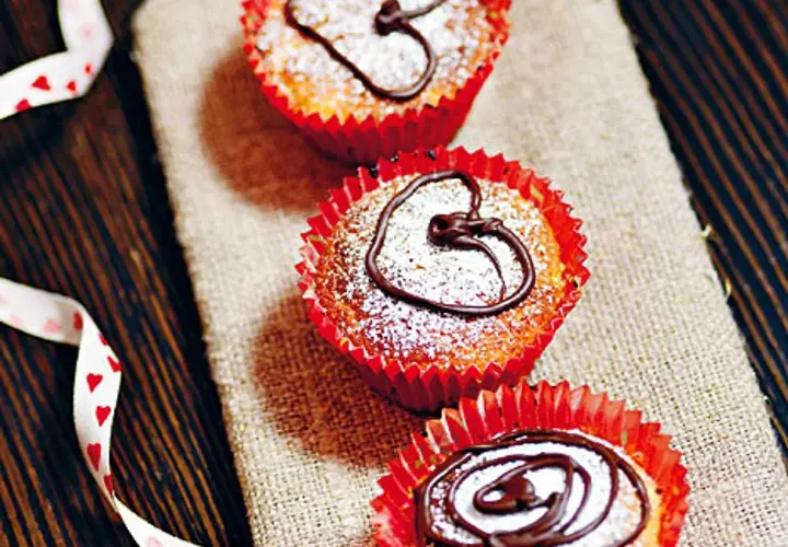 Tre saffransmuffins med mandelmassa, pudrade med florsocker och dekorerade med choklad. En vit sidenband med hjärtan ligger bredvid.