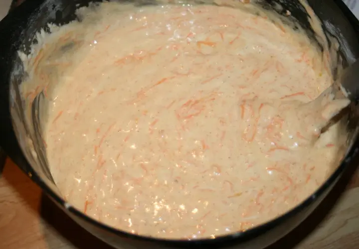 En skål med poröst smör och socker, blandat med rivna morötter. En spatel rör om i smeten.