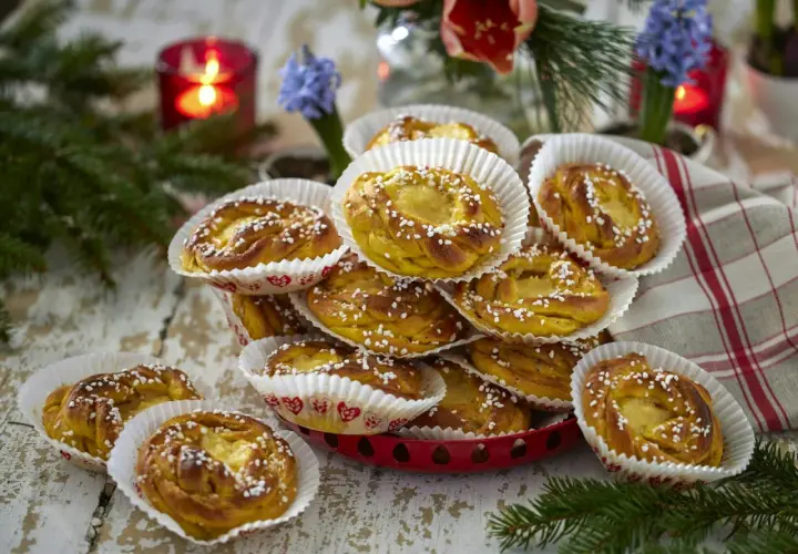 En hög med gyllene kanelbullar, strösslade med pärlsocker, i vita pappersformar. En röd ljusstake och blommor i bakgrunden ger en mysig känsla. Perfekt för att visa hur förvarar man bullar bäst.