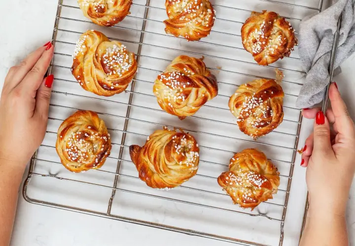 Golden brown cinnamon buns with pearl sugar on a cooling rack, with hands reaching for them.