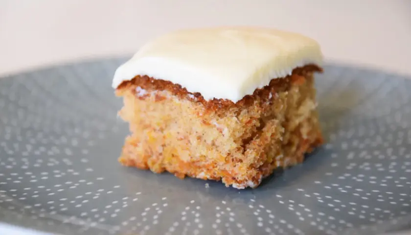 En bit saftig glutenfri morotskaka i långpanna med krämig glasyr på en gråblå tallrik med prickigt mönster.