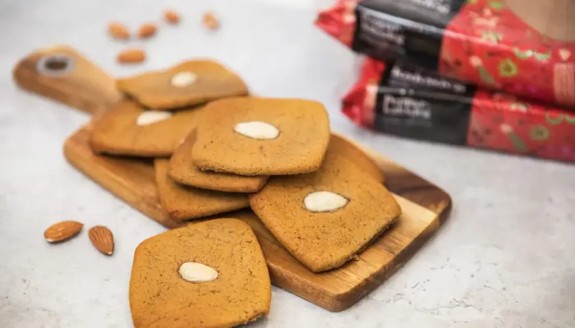 Fler fyrkantiga pepparkakor med mandel ligger på en träskärbräda, med några lösa mandlar och förpackningar i bakgrunden.