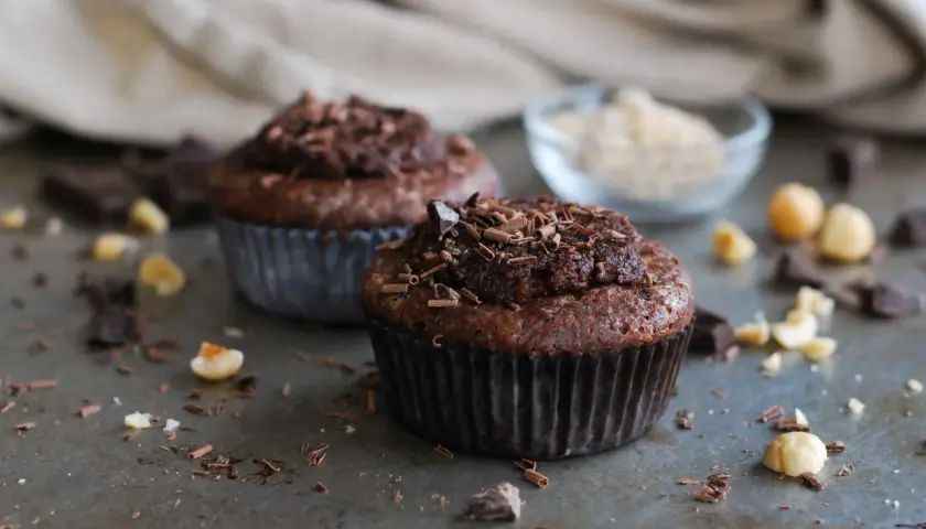 Två chokladmuffins med nutella och strössel av choklad. Hasselnötter och chokladbitar ligger utspridda runt dem.