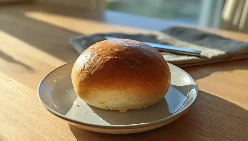 En gyllene, fluffig bulle ligger på en tallrik. Solen lyser in genom fönstret och skapar ett vackert ljus.