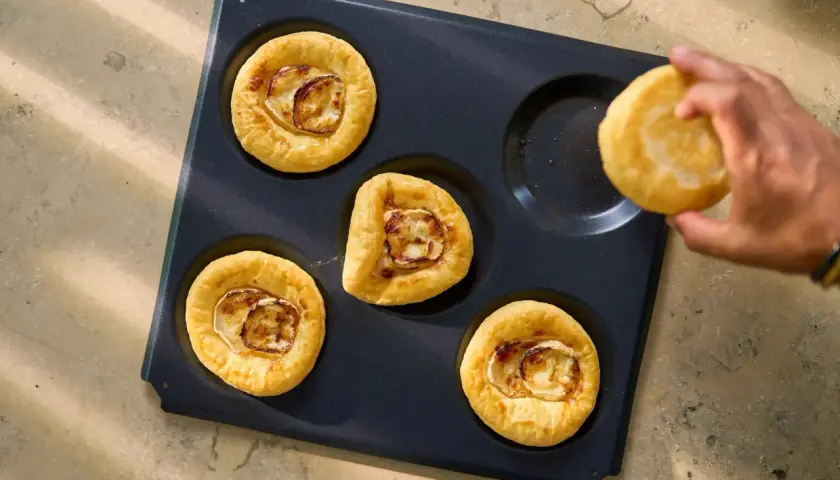 Fyra små pajer med getost och lök ligger i en form, redo att bakas. En hand håller en till paj. Perfekt för att ha två plåtar i ugnen samtidigt.