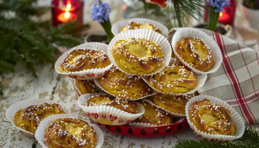 En hög med gyllene kanelbullar, strösslade med pärlsocker, i vita pappersformar. En röd ljusstake och blommor i bakgrunden ger en mysig känsla. Perfekt för att visa hur förvarar man bullar bäst.