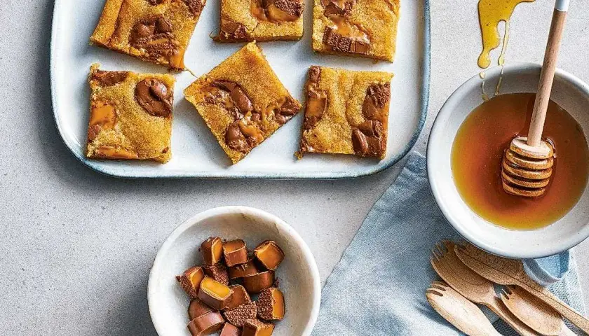Sju bitar av saftiga dumlekakor i långpanna ligger på ett fat. Bredvid finns en skål med honung och en skål med hackade dumlekolor.
