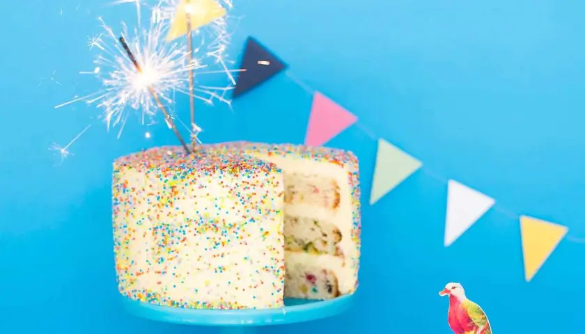 En festlig funfetti tårta med stjärnljus och färgglada girlanger. En liten fågeldekoration sitter bredvid.