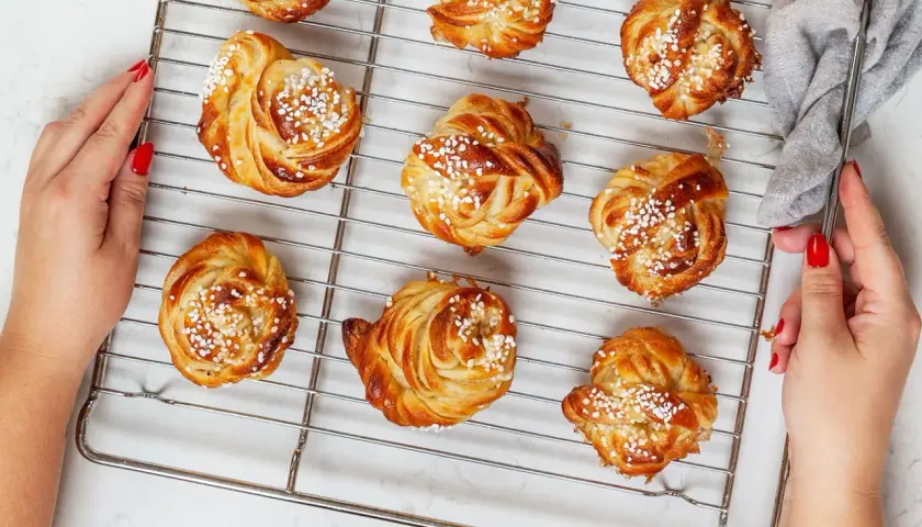 Golden brown cinnamon buns with pearl sugar on a cooling rack, with hands reaching for them.