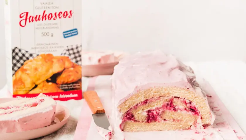En saftig vegansk rulltårta med hallonfyllning och rosa glasyr. Bredvid står en förpackning med glutenfritt mjöl.