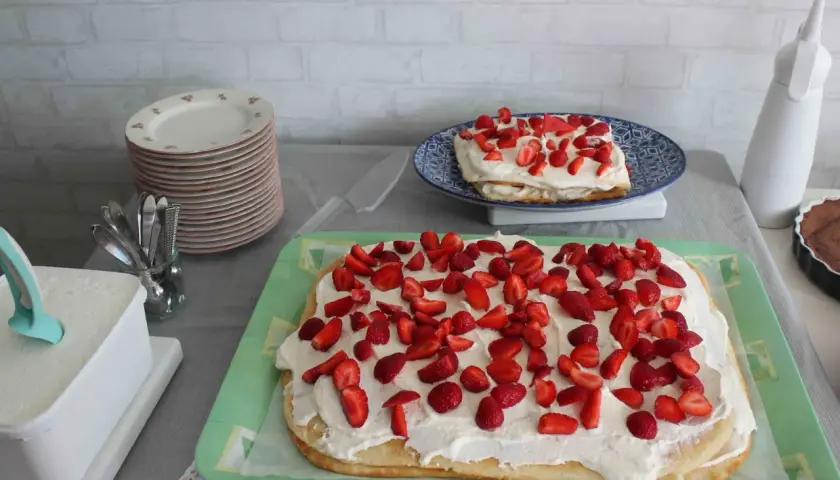 En stor rulltårta som tårta, täckt med grädde och jordgubbar, står på ett grönt fat. En mindre tårta med samma dekoration syns på en blå tallrik.