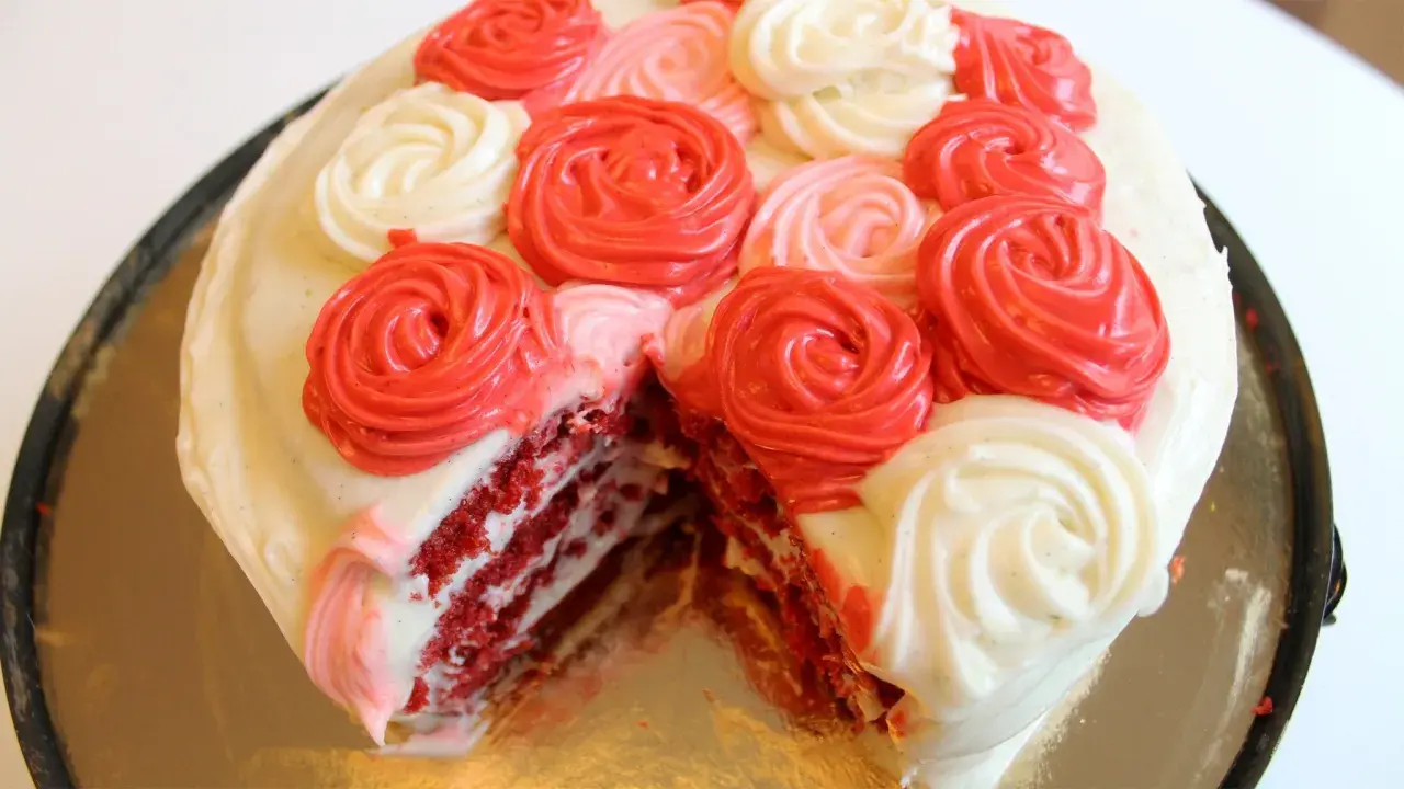 En saftig red velvet-tårta med lager av rött och vitt fluffigt frosting, dekorerad med rosformade frostingblommor i rött och vitt.