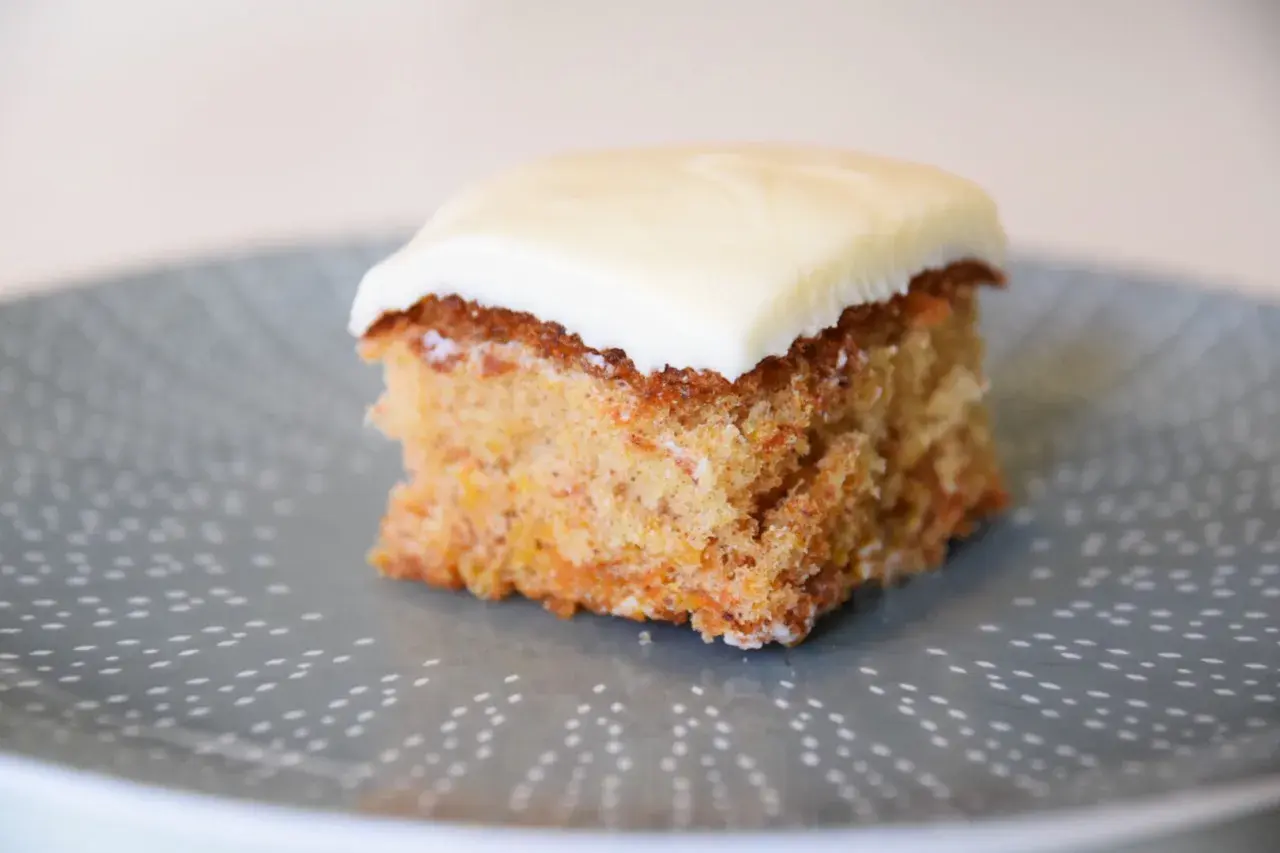 En bit saftig glutenfri morotskaka i långpanna med krämig glasyr på en gråblå tallrik med prickigt mönster.