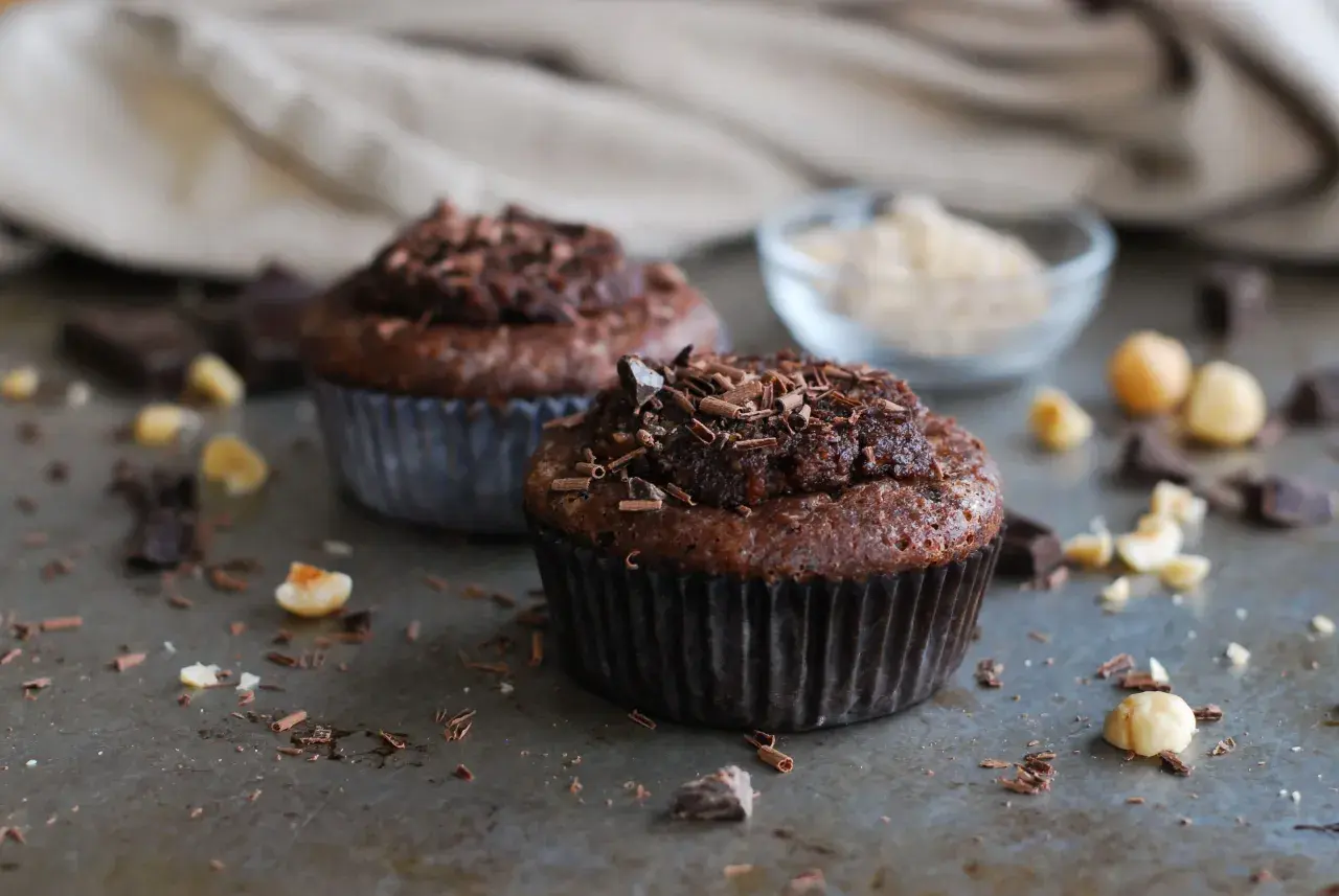 Två chokladmuffins med nutella och strössel av choklad. Hasselnötter och chokladbitar ligger utspridda runt dem.