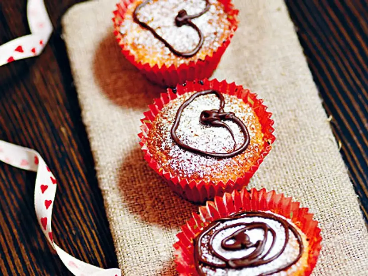 Tre saffransmuffins med mandelmassa, pudrade med florsocker och dekorerade med choklad. En vit sidenband med hjärtan ligger bredvid.