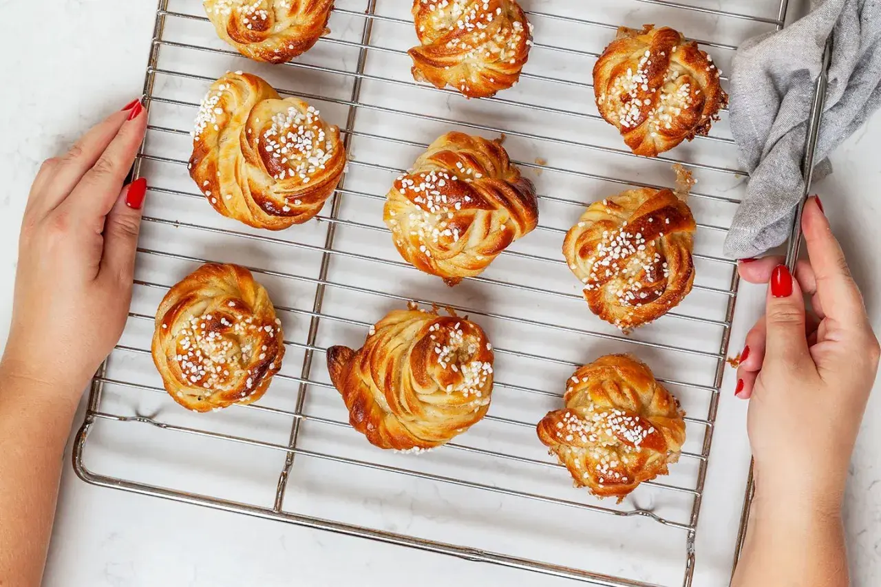 Golden brown cinnamon buns with pearl sugar on a cooling rack, with hands reaching for them.