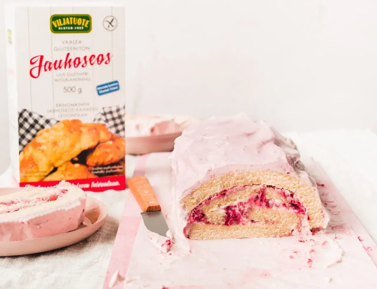 En saftig vegansk rulltårta med hallonfyllning och rosa glasyr. Bredvid står en förpackning med glutenfritt mjöl.
