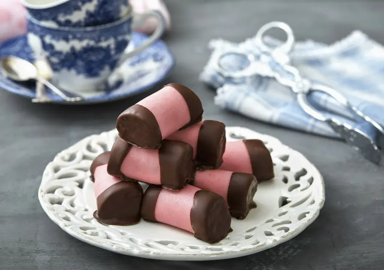 En hög med rosa marsipanbakelser doppade i tempererad choklad, serverade på en vit spetsfat. En blåvit kaffekopp och en duk med tång i bakgrunden.