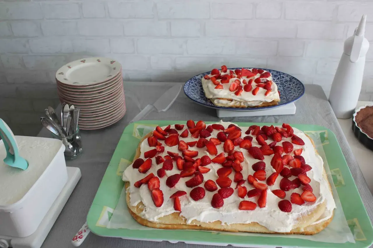 En stor rulltårta som tårta, täckt med grädde och jordgubbar, står på ett grönt fat. En mindre tårta med samma dekoration syns på en blå tallrik.