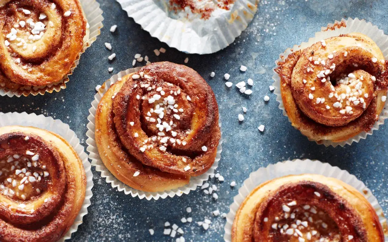 Flera kanelbullar med pärlsocker ligger på ett blått underlag. Kanelbulle degens gyllene färg och sockerströssel ser aptitligt ut.