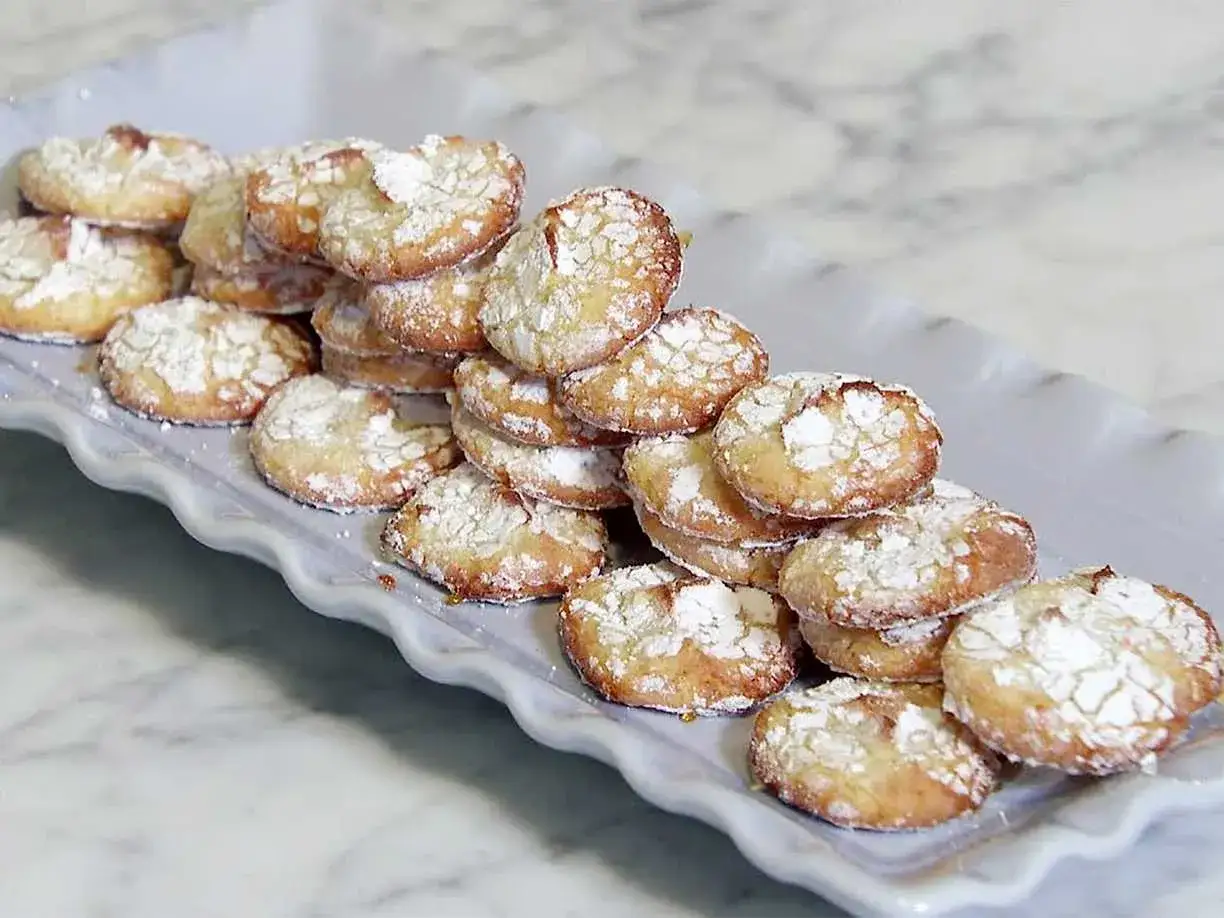 En hög med hembakade amaretti-kakor, pudrade med florsocker, på ett vitt fat. Ett perfekt amaretti recept!