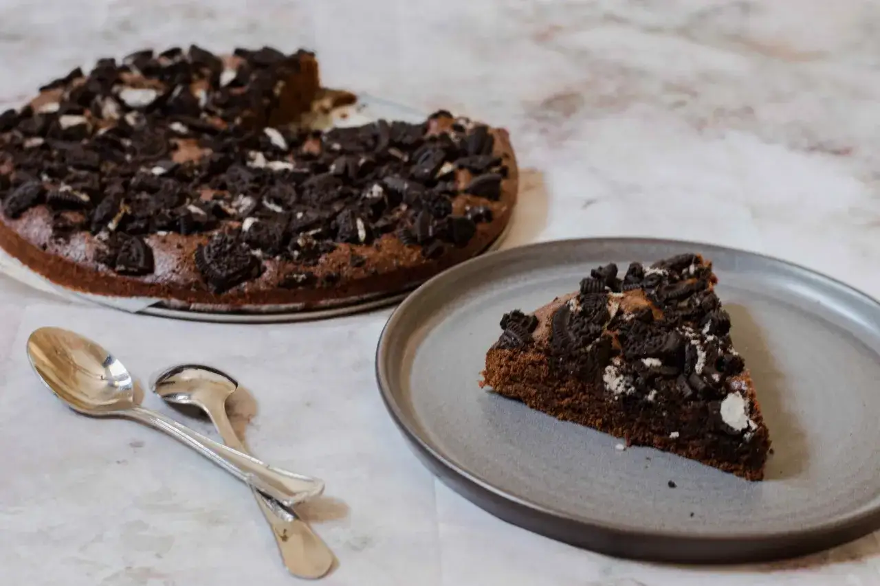 En bit chokladig oreo kladdkaka p&aring; en tallrik, med en hel kaka i bakgrunden. Tv&aring; skedar ligger bredvid.