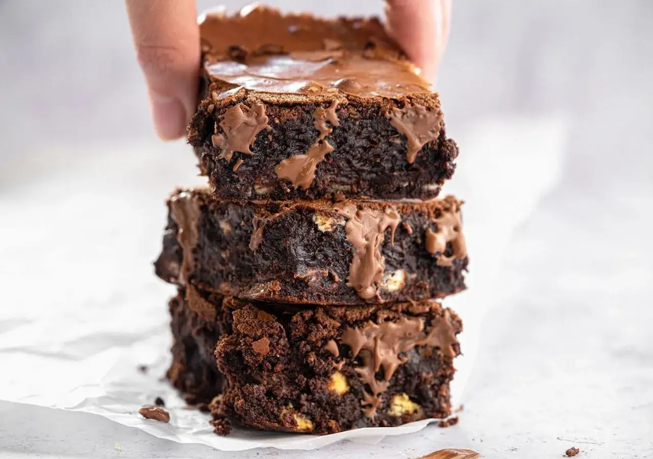 En hand lyfter en stapel saftiga, mörka glutenfria brownies med smält choklad.