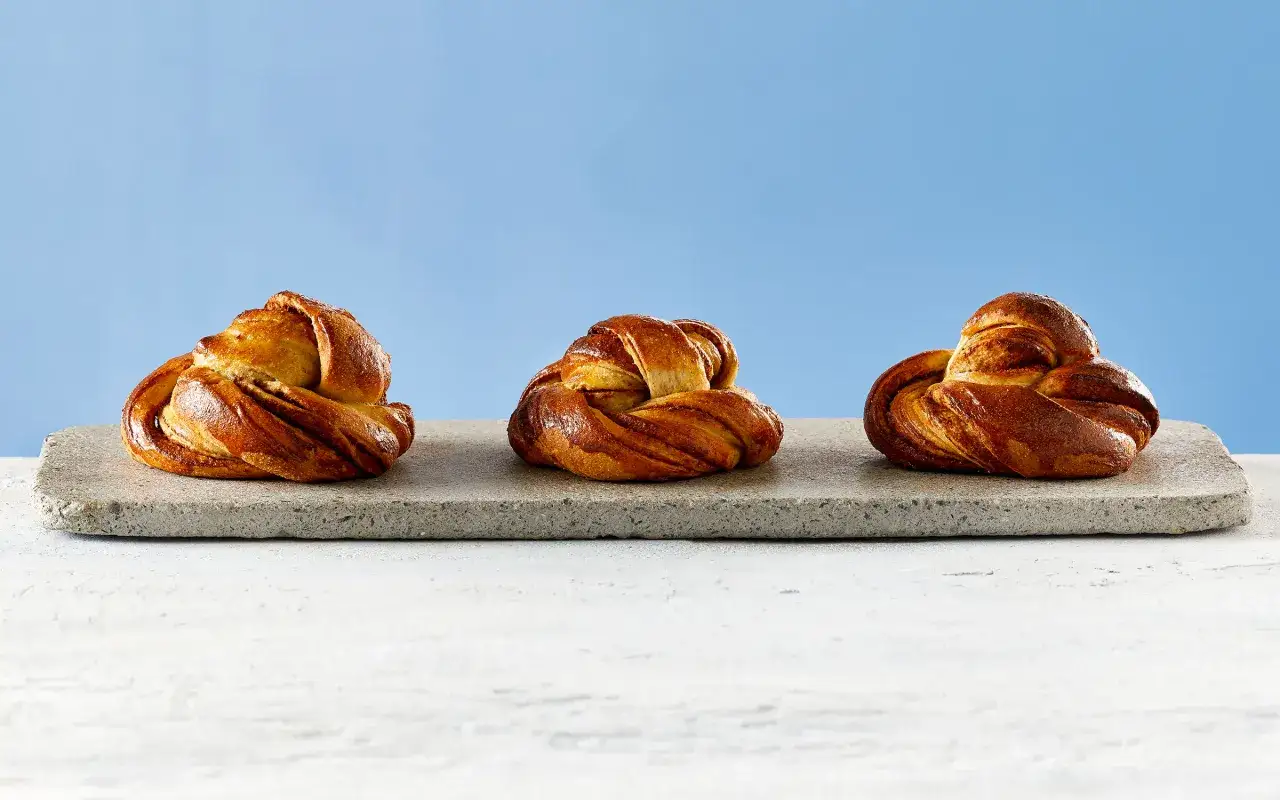 Tre gyllene laktosfria kanelbullar ligger på en stenplatta mot en blå bakgrund.