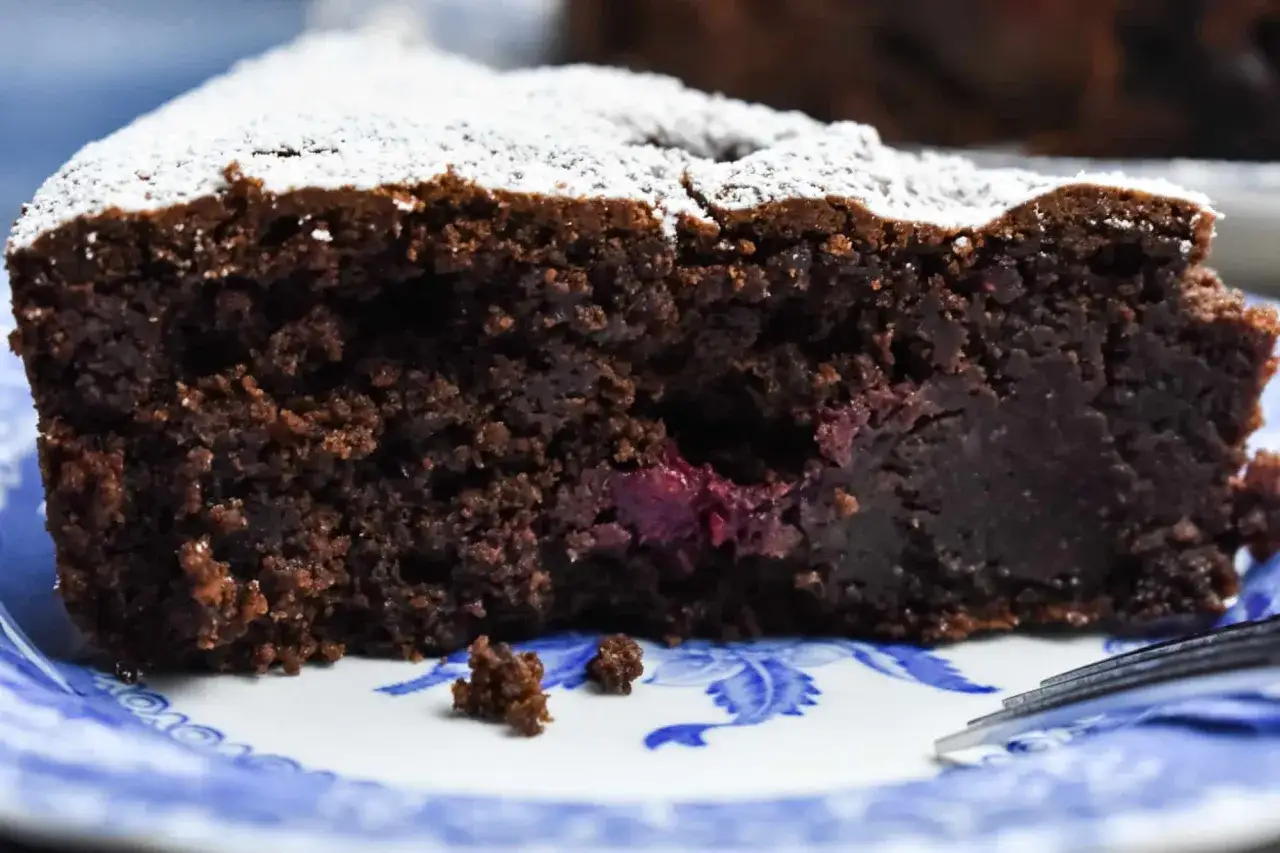 En saftig bit glutenfri chokladkaka med körsbär, pudrad med florsocker, serverad på en blåvit tallrik.