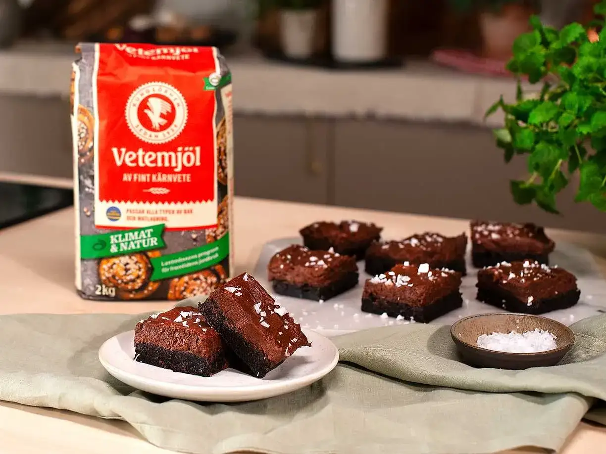 M&ouml;rka, fudgiga chokladbrownies toppade med flingsalt, redo att avnjutas. Bredvid st&aring;r en p&aring;se vetemj&ouml;l, ingrediensen som g&ouml;r dessa chokladbrownies m&ouml;jliga.