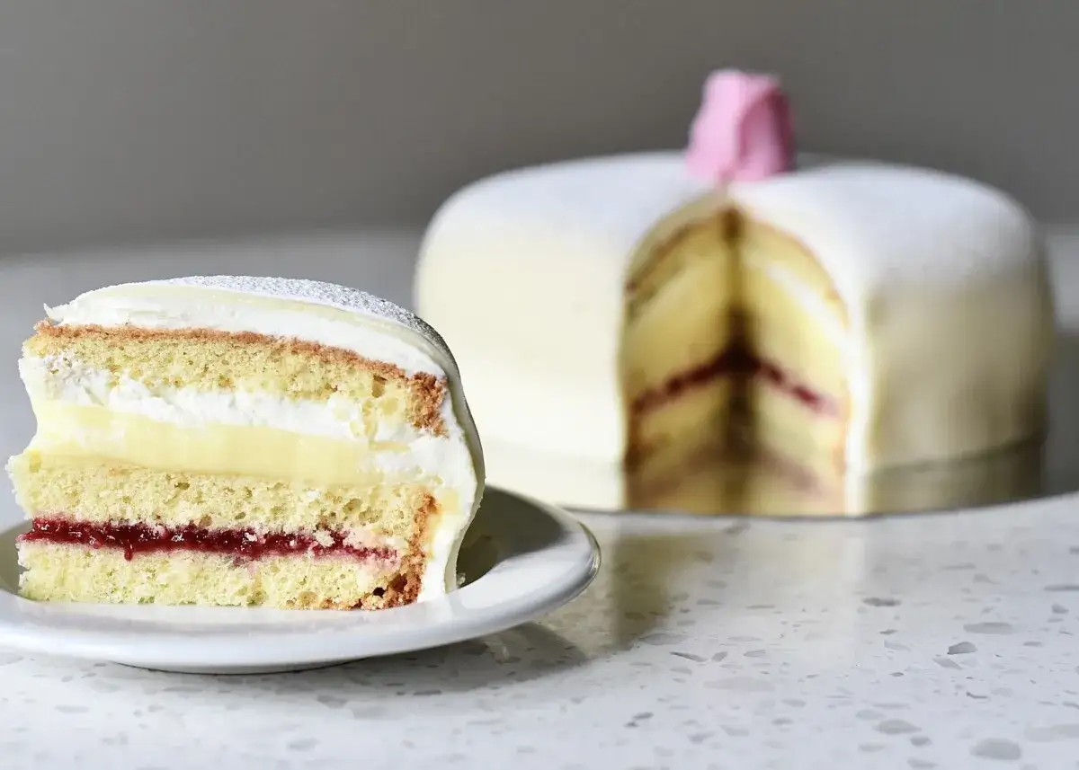 En skiva av en vacker butterkaka med vaniljkr&auml;m och hallonsylt. Hela t&aring;rtan syns i bakgrunden, dekorerad med en rosa ros.