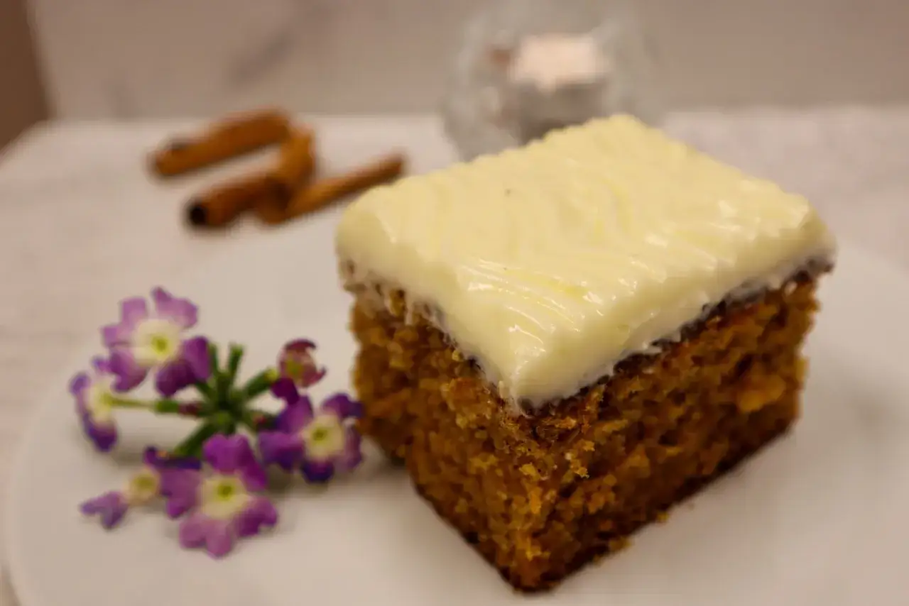 En bit glutenfri morotskaka i l&aring;ngpanna med kr&auml;mig frosting, dekorerad med lila blommor och kanelst&auml;nger i bakgrunden.