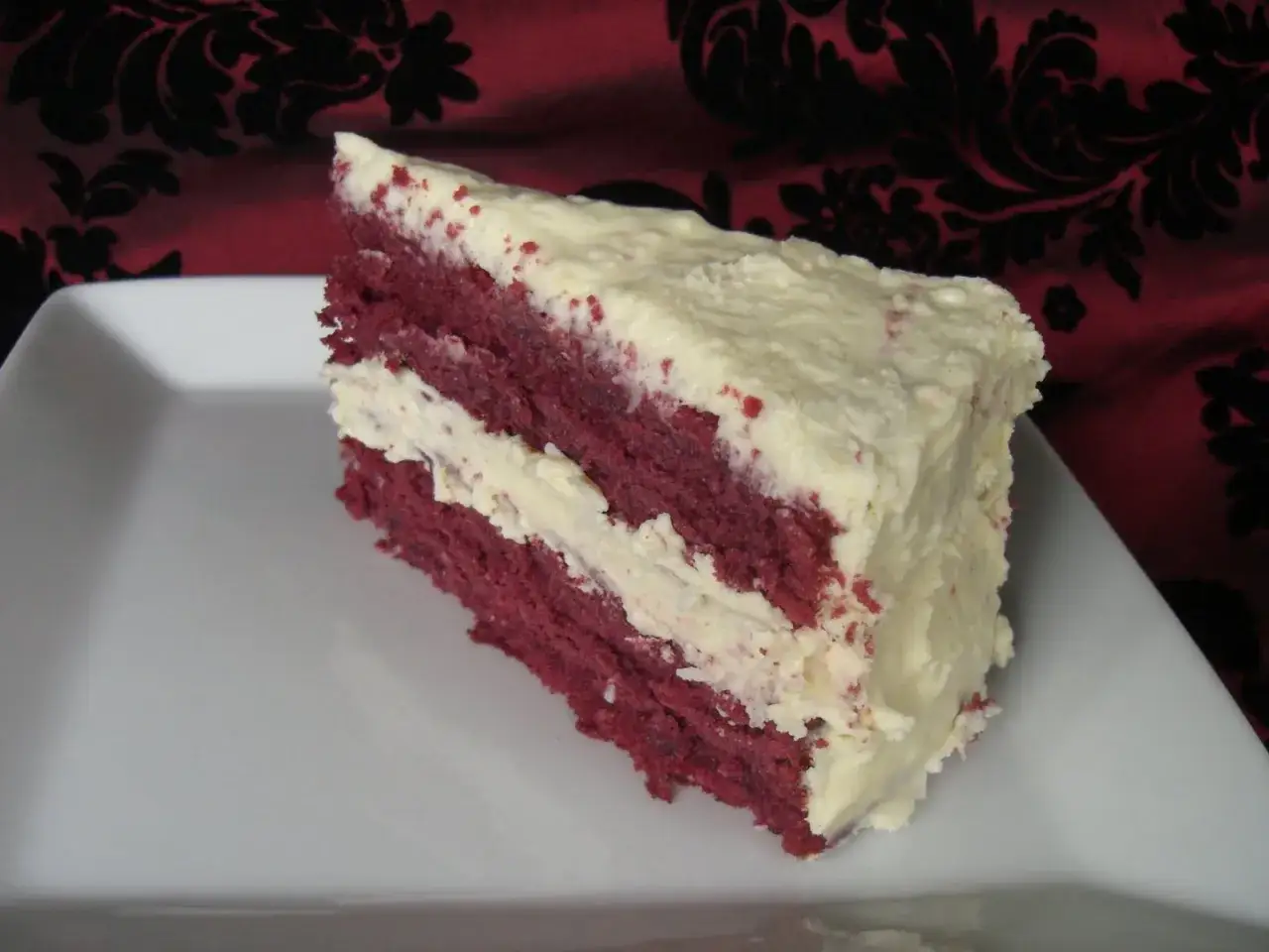 En bit saftig red velvet-tårta med krämig frosting på en vit tallrik.