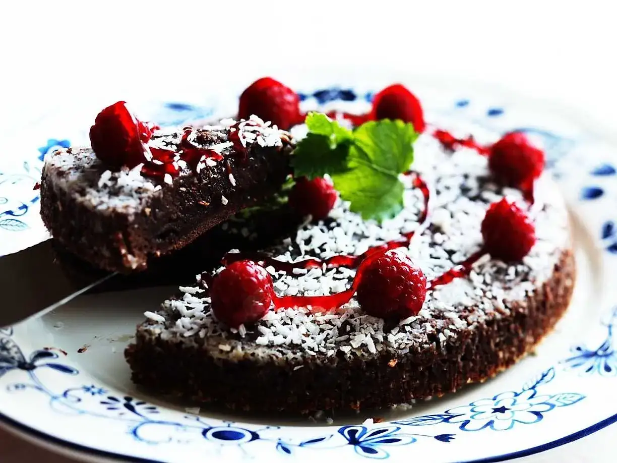 En saftig kladdkaka med choklad, toppad med kokosflingor, hallon och en röd sås. En bit är uppskuren och redo att avnjutas.