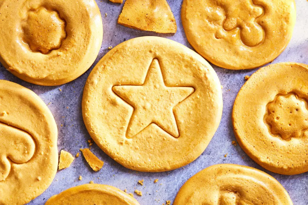 Flera gyllene dalgona cookies med olika former, som en stj&auml;rna och en blomma, ligger utspridda p&aring; en gr&aring; yta.