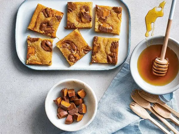 Sju bitar av saftiga dumlekakor i långpanna ligger på ett fat. Bredvid finns en skål med honung och en skål med hackade dumlekolor.
